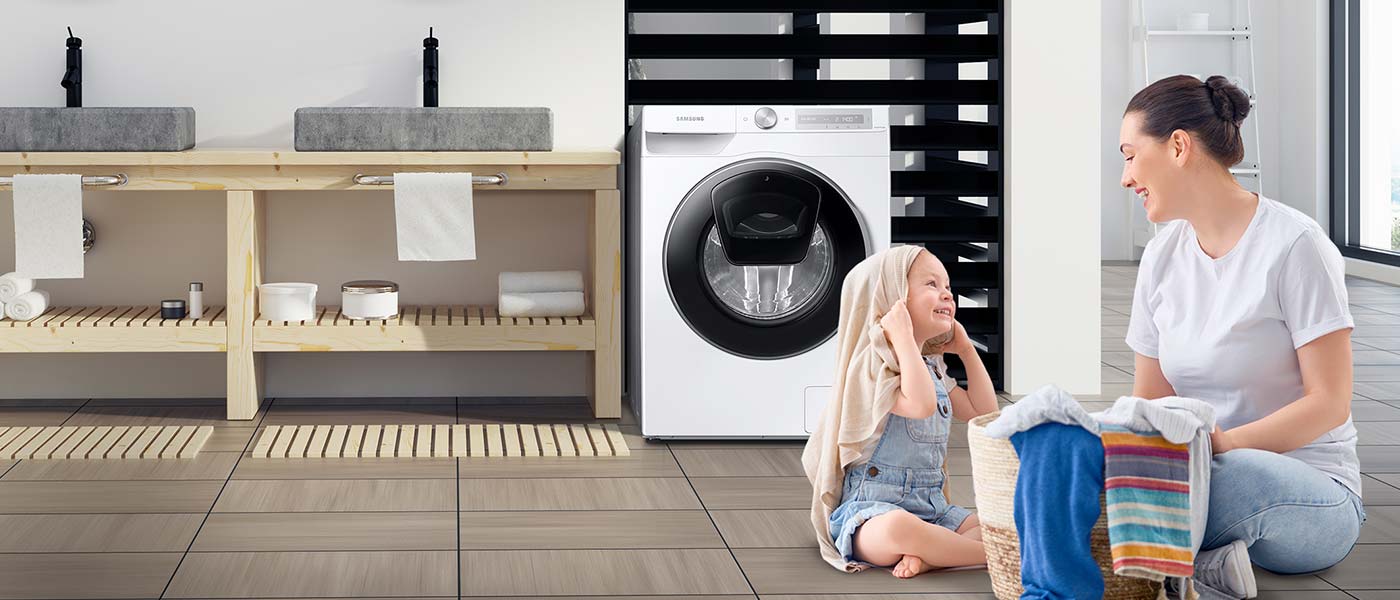 Усміхаючись жінка з дівчинкою, дитина грає вранці, пральна машина Samsung на задньому плані.