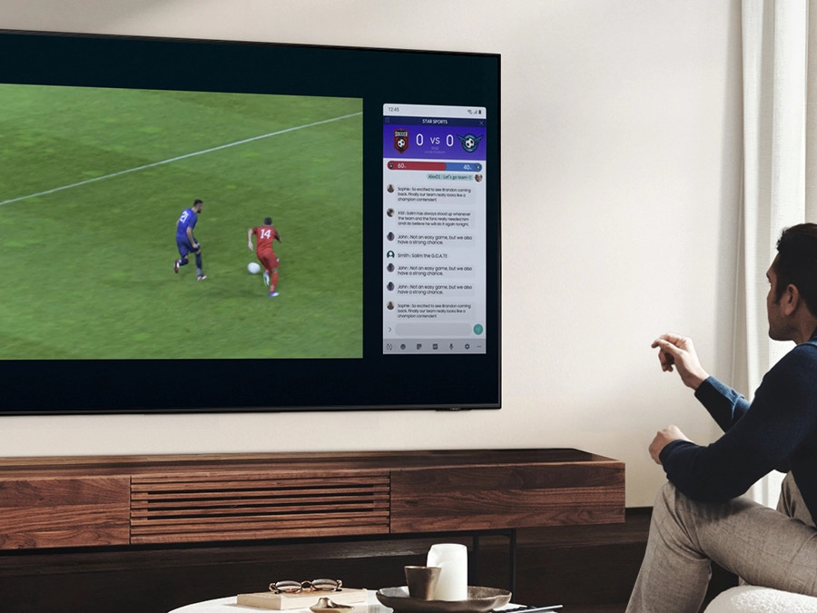 Мужчина использует QLED Multi View, чтобы одновременно смотреть футбольный матч и читать новости на одном экране.  Q80BATXXH