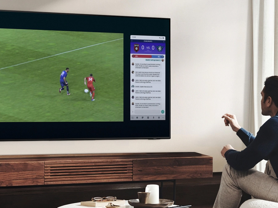 Мужчина использует QLED Multi View, чтобы одновременно смотреть футбольный матч и читать новости на одном экране.