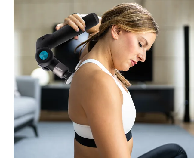 Жінка в короткому білому топі з чорним шнурком масажує верхню частину спини масажним пістолетом Raven, на задньому плані видно кімнату.