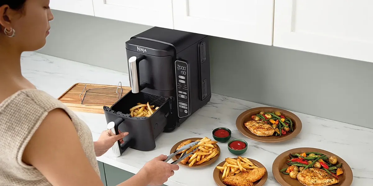 Людина викладає картоплю фрі з аерофритюрниці Ninja на тарілки з куркою та овочами на кухонному столі.