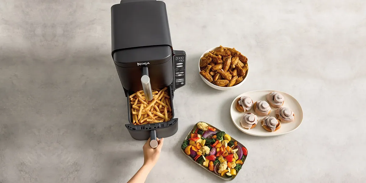 Аерофритюрниця Ninja з відкритою шухлядою, повною картоплі фрі, поруч із мискою зі стравами: крильцями, овочами та булочками