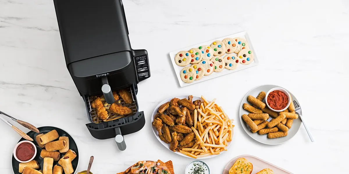 Аерофритюрниця Ninja з відкритою шухлядою з нагетсами, поруч з тарілками з картоплею фрі, крильцями, закусками та печивом