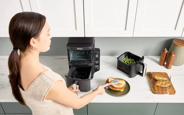 Жінка кладе тост з аерофритюрниці Ninja на тарілку; на стільниці поруч лежить шухляда з печеною квасолею та дошка хліба.
