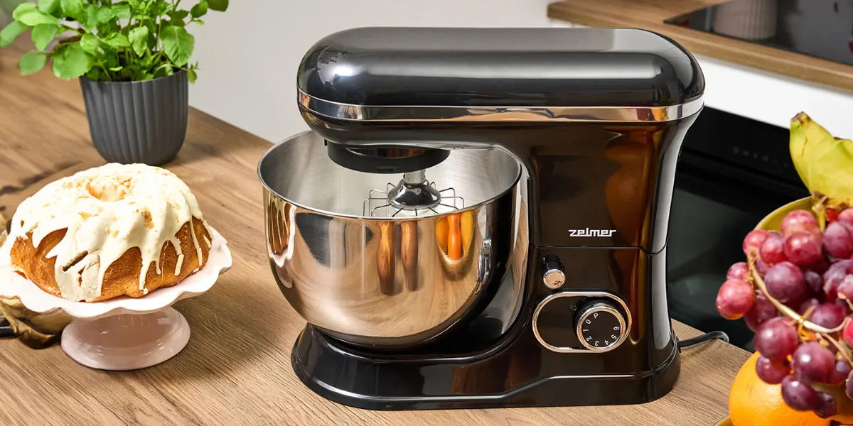 Чорний планетарний кухонний комбайн Zelmer з металевою чашею на кухонній стільниці, поруч з ним тісто, трави та фрукти.