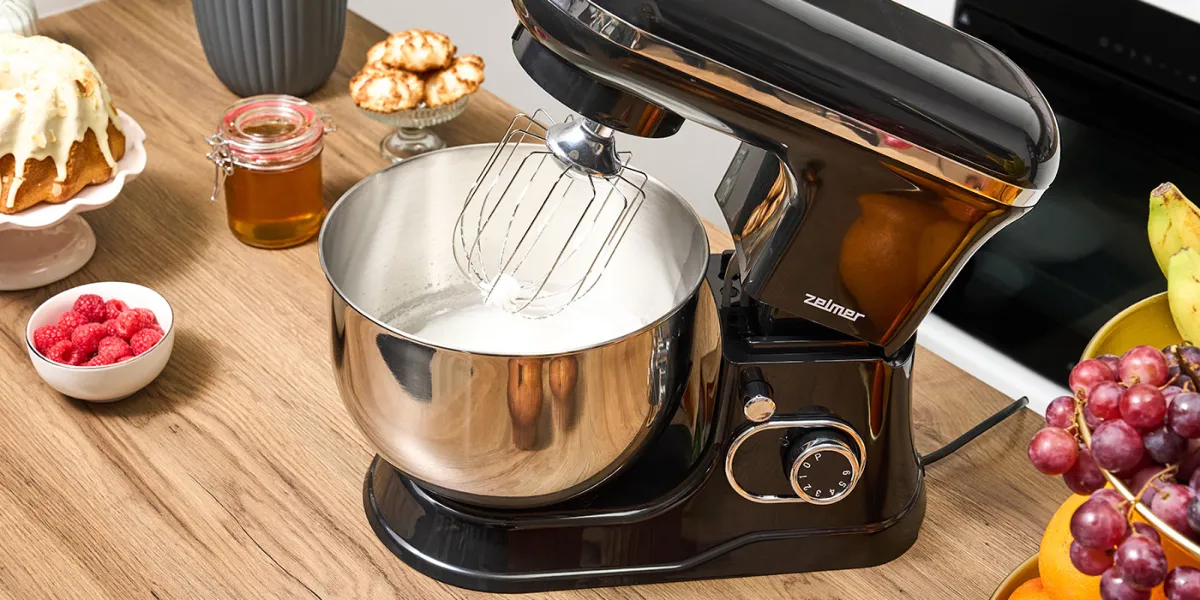 Планетарний міксер Zelmer з металевою чашею та вінчиком на кухонній стільниці, поруч із ним — тісто, фрукти та інгредієнти для випічки.