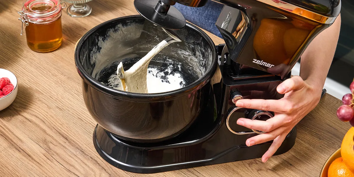 Людина вмикає чорний планетарний кухонний комбайн Zelmer з гаком для тіста, і в чаші видно дріжджове тісто.