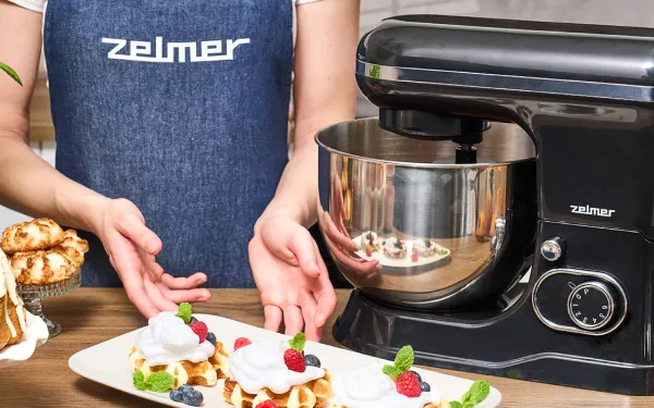 Людина у фартусі Zelmer презентує вафлі з фруктами, поруч — чорний планетарний робот Zelmer з металевою мискою.