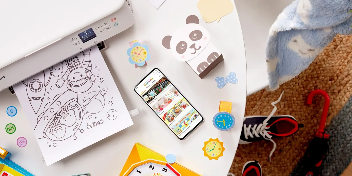 Принтер Canon PIXMA друкує розмальовку, навколо розставлені іграшки, а на столі лежить смартфон.