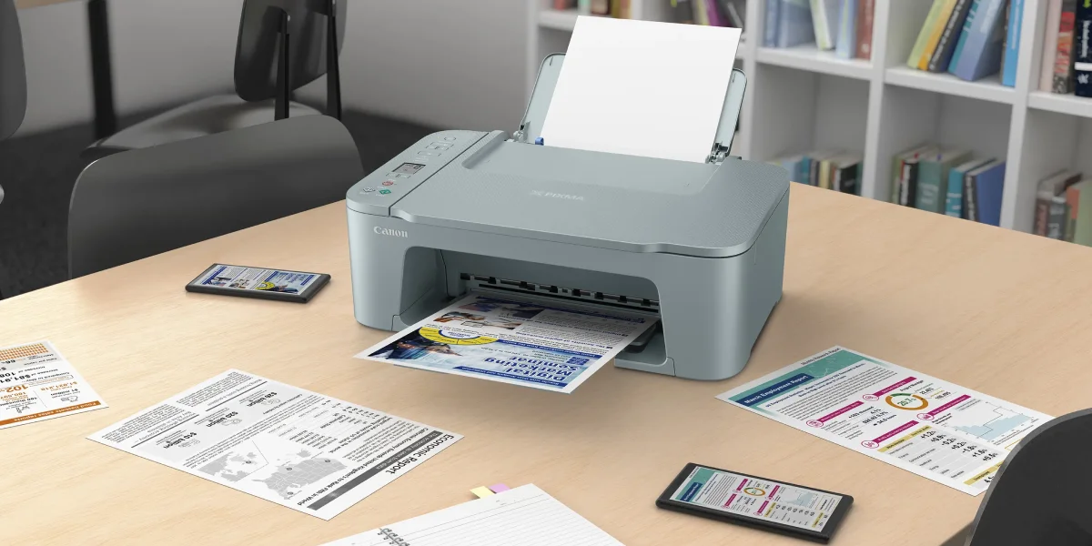 Drukarka Canon na biurku podczas drukowania kolorowej strony, otoczona smartfonami i dokumentami
