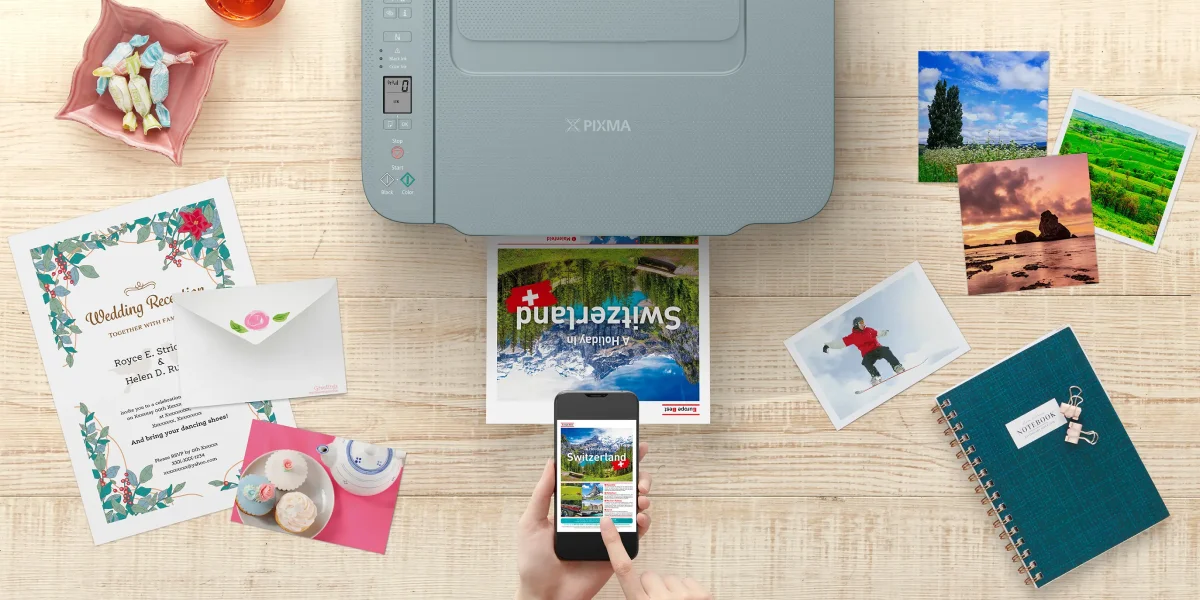 Widok z g&oacute;ry: drukarka Canon drukuje zdjęcie Szwajcarii z aplikacji mobilnej, otoczona zdjęciami i zaproszeniem