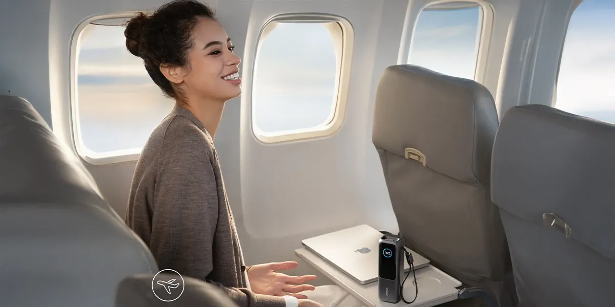 Усміхнена жінка в літаку поруч із портативним зарядним пристроєм Anker, екран якого показує 100% заряду батареї.