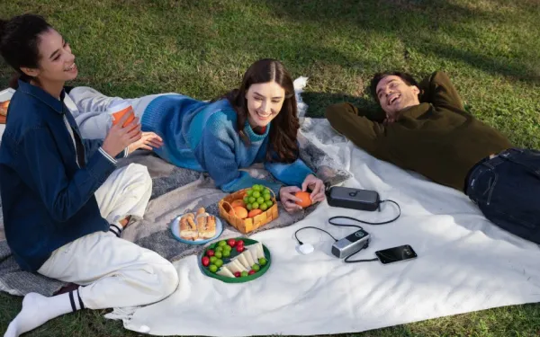 Троє людей пікнікують на траві. Портативний зарядний пристрій Anker заряджає телефон та інші пристрої.
