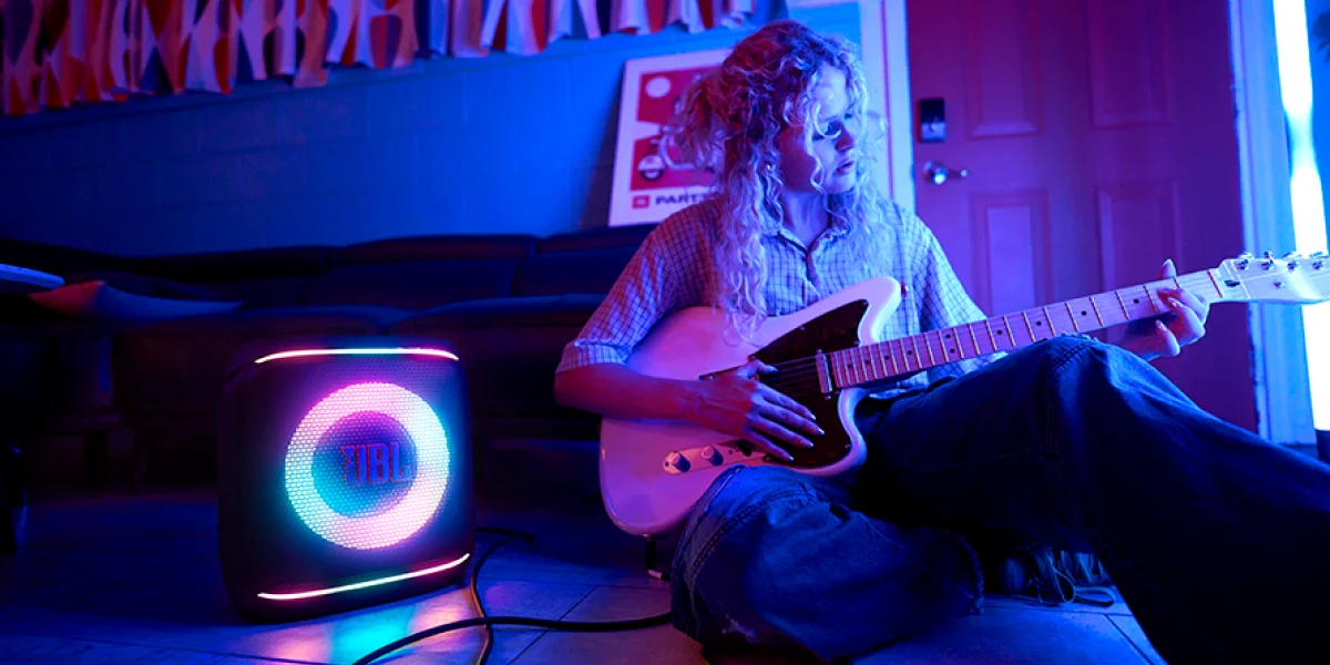 Людина сидить на підлозі та грає на гітарі поруч із динаміком JBL із синім та рожевим підсвічуванням.