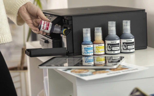Людина додає рожеве чорнило до принтера Brother, поруч стоять пляшечки з чорнилом інших кольорів.
