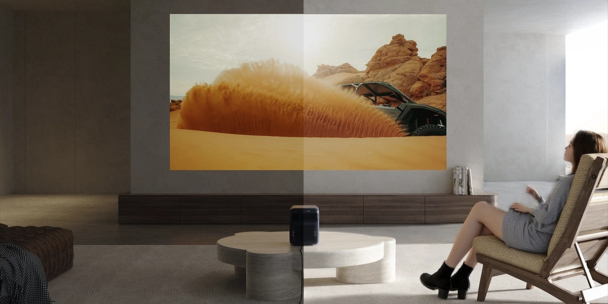 Проектор проектує зображення пустельного транспортного засобу на стіну, а навпроти нього у сучасній, світлій вітальні сидить жінка.