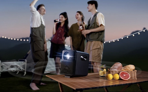 Проектор світить на стіл під час вечірнього кемпінгу, поки група друзів п'є та базікає на задньому плані.