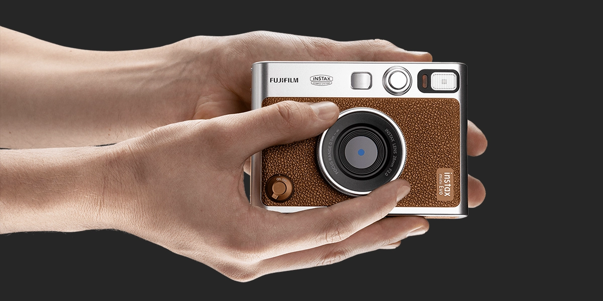 Дві руки тримають коричневу камеру Instax. Пальці повертають срібне кільце на об'єктиві, яке змінює ефекти.