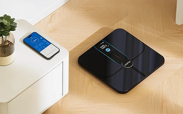 Ваги Renpho Elis Nova лежать на підлозі, а на шафі — смартфон із фітнес-додатком.