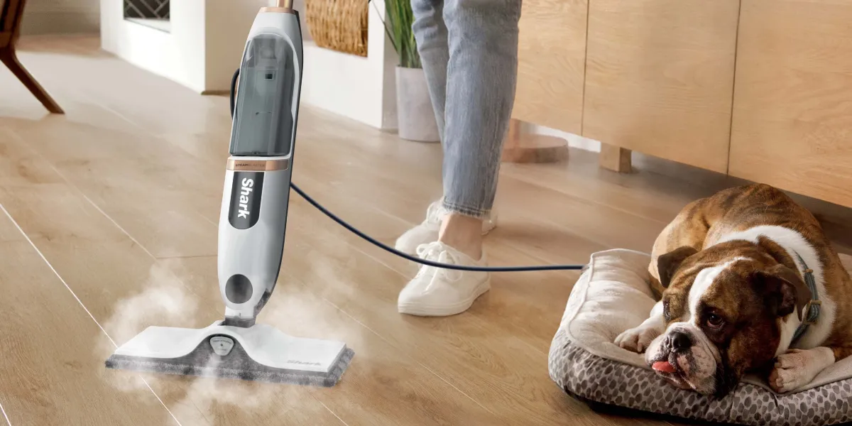 Людина чистить дерев'яну підлогу паровою шваброю Shark S2001EU, поруч із собакою, що лежить на ліжку