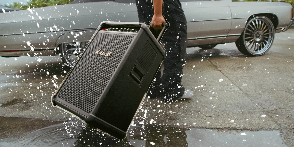 Людина тягне колонку Marshall на колесах через калюжу поруч із припаркованим автомобілем.